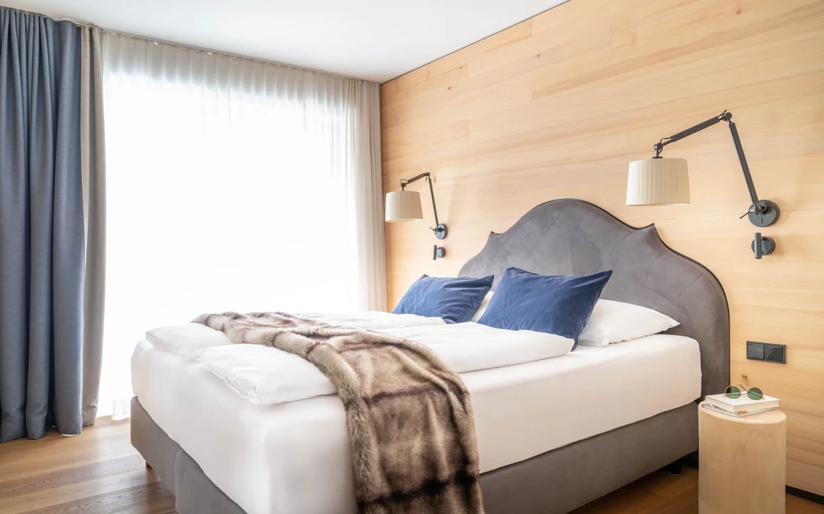 Ein großes Bett mit grauer Polsterung, einer weißen Decke und einem blauen Vorhang im Hintergrund | © Silvretta Montafon - blateral