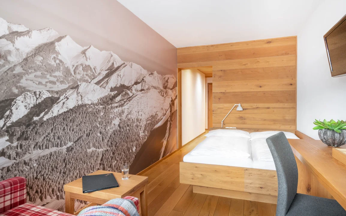 Ein Doppelbett mit Holzrahmen vor einer Holzwand und eine rot-karierter Couch | © Silvretta Montafon - blateral