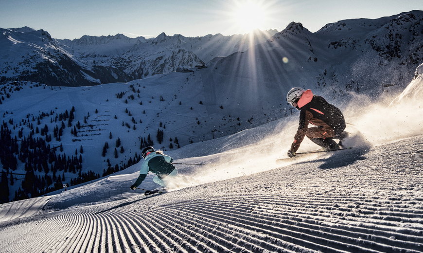 Zwei Personen in Skikleidung und Helm sausen frisch präparierte Pisten mit Bergpanorama im Hintergrund hinunter