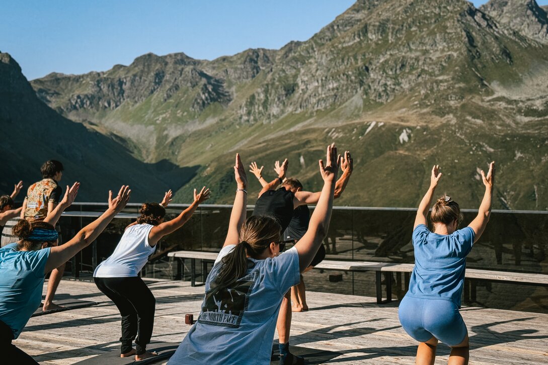 Eine Gruppe von Personen steht auf einer Terrasse bei Morgensonne und macht Yoga. | © Silvretta Montafon - Vanessa Strauch