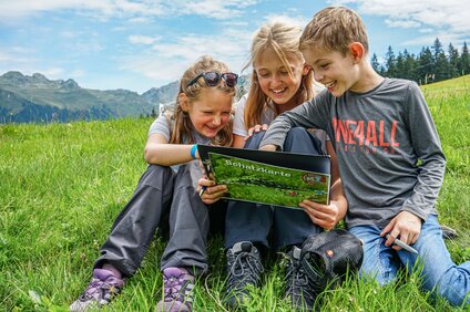 Drei Kinder sitzen auf einer Wiese und lesen eine Schatzkarte in der Silvretta Montafon. | © Silvretta Montafon - Vanessa Strauch