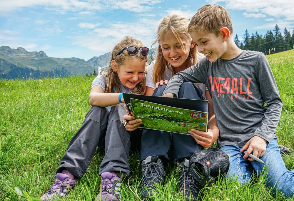 Schatzsuche - Silvretta Montafon | © Silvretta Montafon - Vanessa Strauch Drei Kinder sitzen auf einer Wiese und lesen eine Schatzkarte in der Silvretta Montafon. | © Silvretta Montafon - Vanessa Strauch