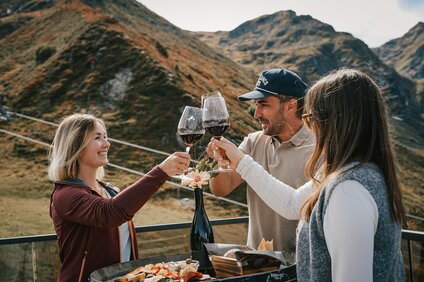 Drei Personen stehen an einem Weinfass mit Käse und Brot und stoßen mit einem Glas Rotwein an. | © Silvretta Montafon - Vanessa Strauch
