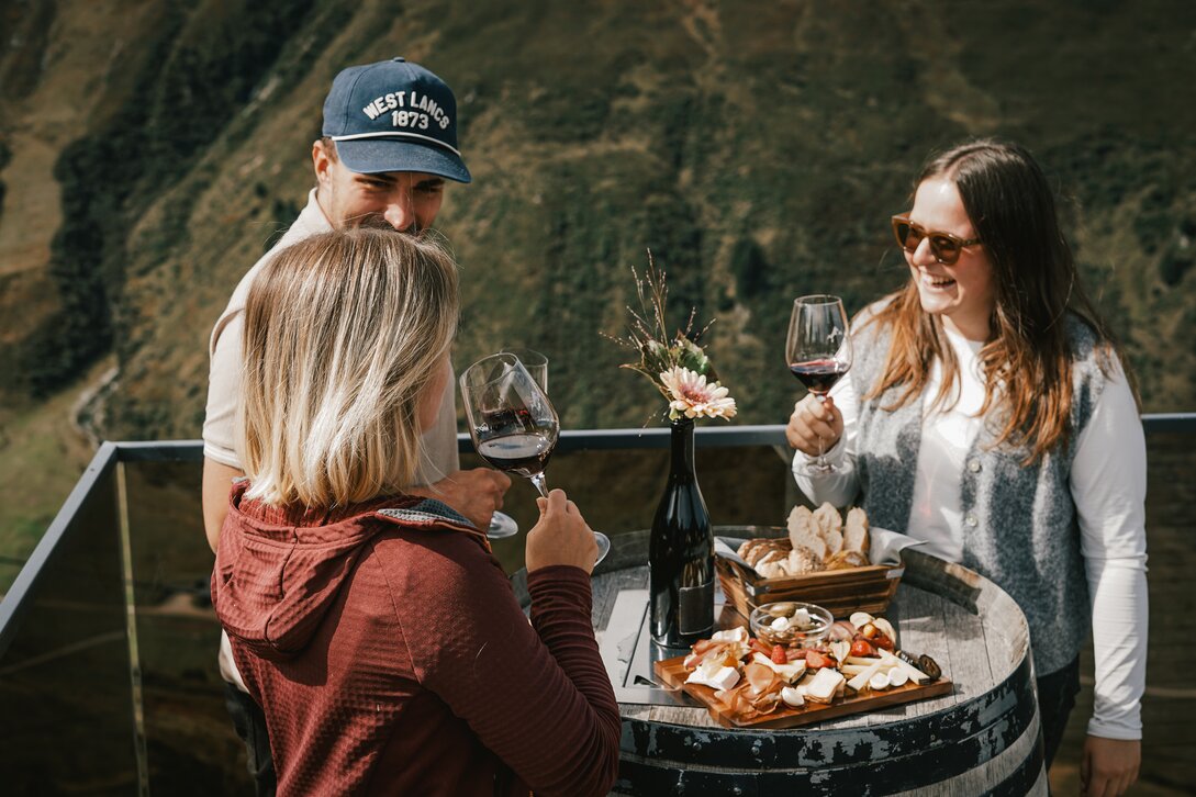 Drei Personen stehen an einem Weinfass mit Käse und Brot und genießen ein Glas Rotwein. | © Silvretta Montafon - Vanessa Strauch
