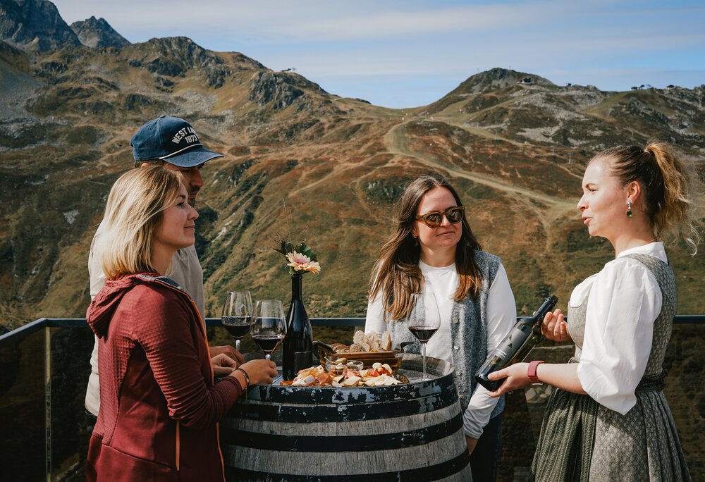 Weinprobe - Silvretta Montafon | © Silvretta Montafon - Vanessa Strauch Drei Personen stehen an einem Weinfass mit einem Glas Rotwein an und eine Kellerin im Dirndl erklärt den Wein. | © Silvretta Montafon - Vanessa Strauch