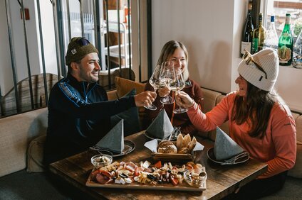 Drei Personen an einem Tisch mit Käse und Brot sitzen in einer Weinstube und stoßen mit einem Glas Weißwein an. | © Silvretta Montafon - Vanessa Strauch