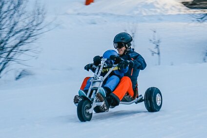 Ein Mann mit einem Kind in Skikleidung auf einem Mountaincart auf der verschneiten Piste. | © Silvretta Montafon - Vanessa Strauch