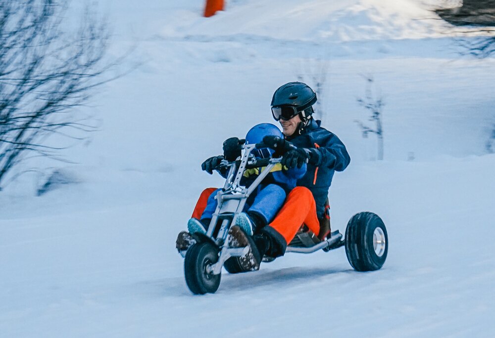 Ein Mann mit einem Kind in Skikleidung auf einem Mountaincart auf der verschneiten Piste. | © Silvretta Montafon - Vanessa Strauch