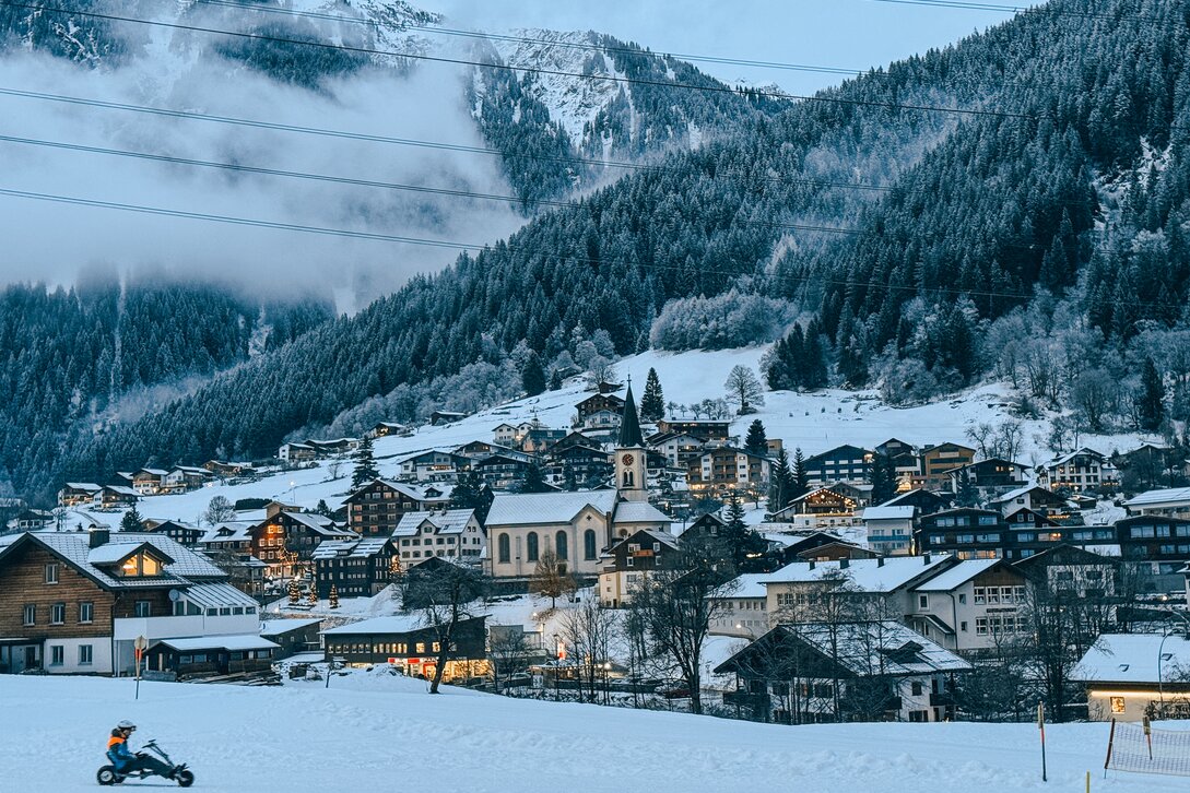 Eine Person in Skikleidung auf der Mountaincarts-Strecke, das verschneite Dorf Gaschurn im Abendlicht im Hintergrund. | © Silvretta Montafon - Vanessa Strauch