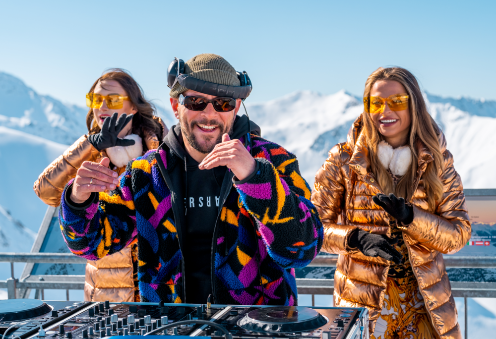 Sebastian Bronk - Silvretta Montafon | © Silvretta Montafon - Sebastian Bronk Sebastian Bronk am DJ Pult mit zwei Tänzerinnen in Orange im Hintergrund | © Silvretta Montafon - Sebastian Bronk