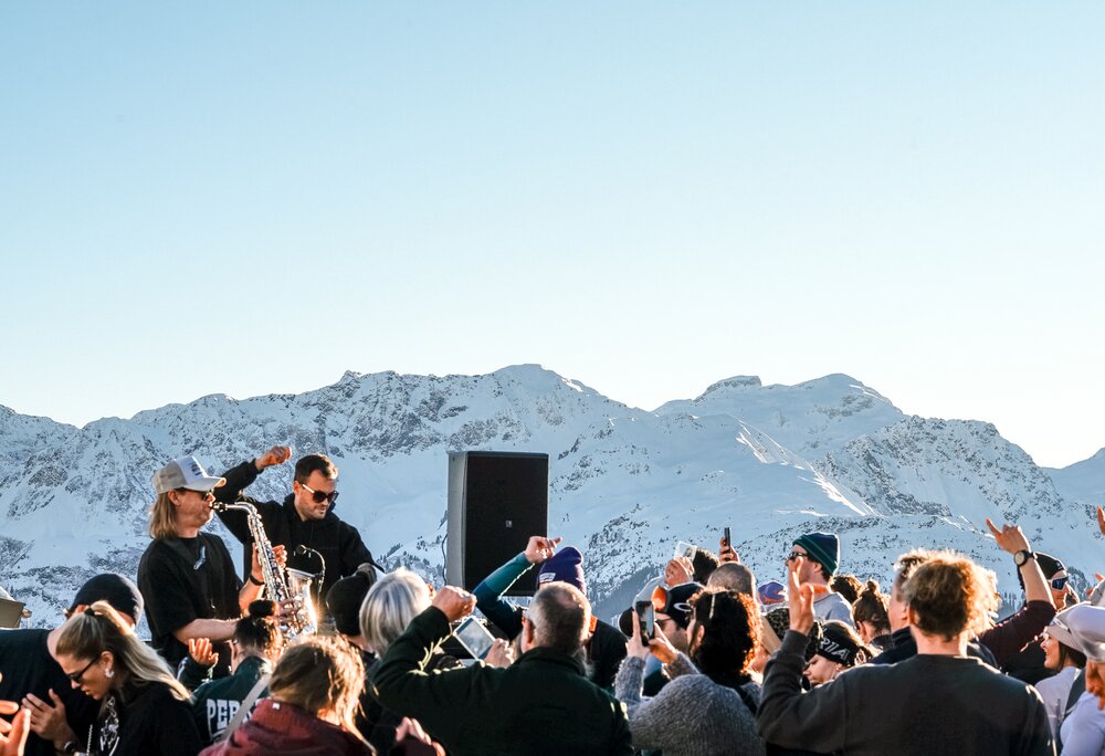 Zwei Musiker, einer mit Saxophon auf einer Terrasse mit vielen Gästen bei Sonnenuntergang und Panorama auf die verschneiten Gipfel. | © Silvretta Montafon - Vanessa Strauch