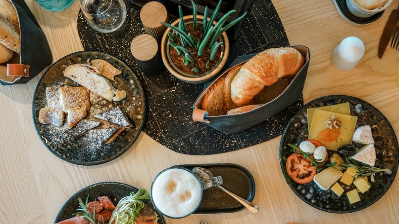 Ein reichlich gedeckter Frühstückstisch mit Milchkaffee, Brotkorb, Wurst, Käse, süßem Gebäck und Frühstücksei. | © Silvretta Montafon - Vanessa Strauch