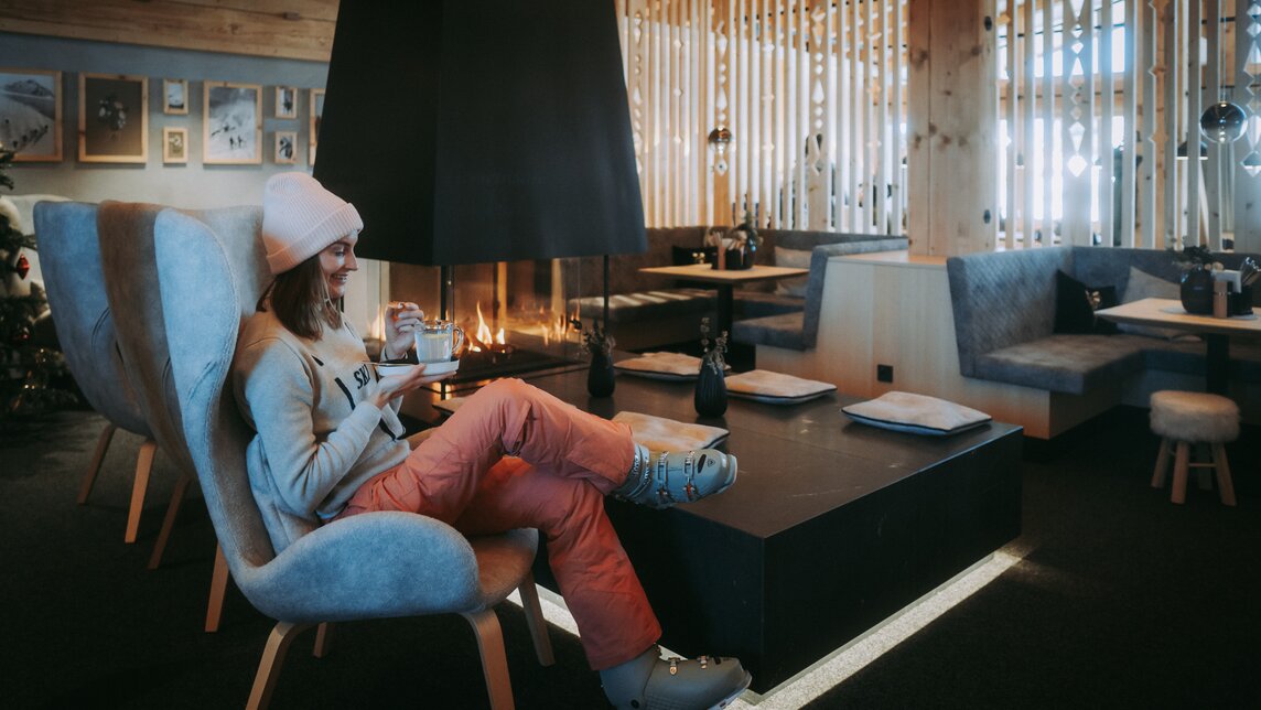 Eine Frau wärmt sich in Skikleidung mit Kaffee in der Hand an einem Kamin im Bergrestaurant. | © Silvretta Montafon - WanderlustAllgäu