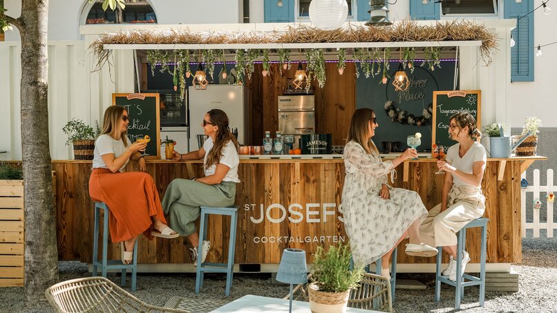 Vier Frauen in bunter Sommerkleidung sitzen an der Bar von St. Josefs Cocktailbar und schlürfen Cocktails | © Silvretta Montafon - Vanessa Strauch