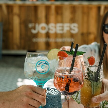 Vier unterschiedliche Cocktails stoßen an mit einer Frau mit Sonnenbrille im Hintergrund | © Silvretta Montafon - Vanessa Strauch