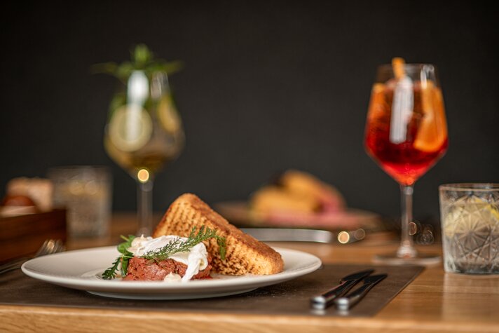 Ein Teller mit Beef Tartar mit Getränken im Hintergrund in Simons Alpenbrasserie. | © Silvretta Montafon - Mara Baues