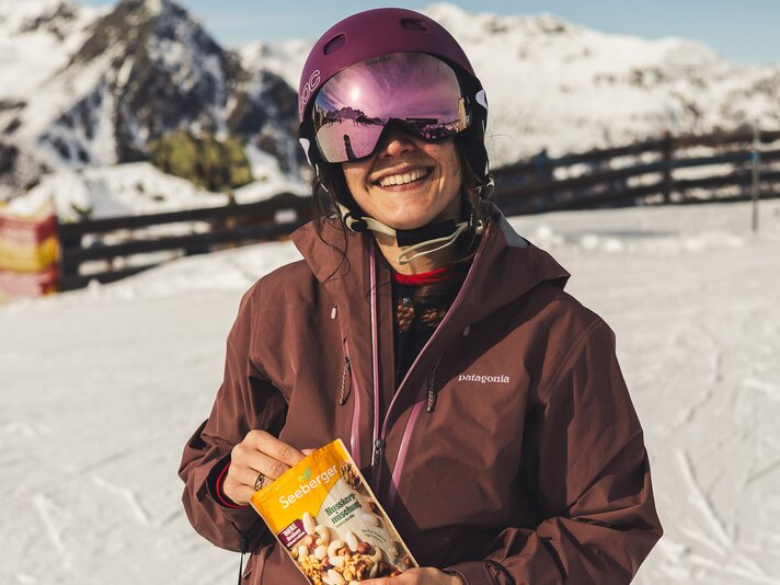 Eine Fraue lacht mit Skikleidung und Skibrille in die Kamera und hält eine Packung Nüsse in der Hand. | © Silvretta Montafon - Roadtyping