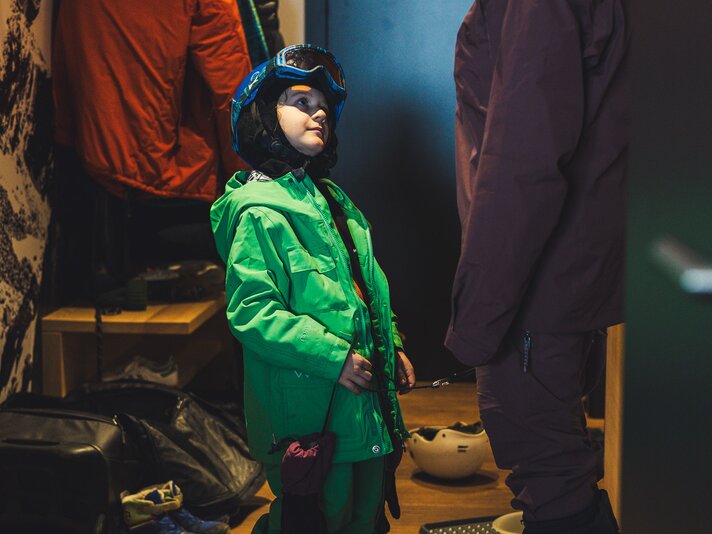 Eine Junge in Skikleidung und Helm schaut seine Mutter im Hotelzimmer an. | © Silvretta Montafon - Roadtyping
