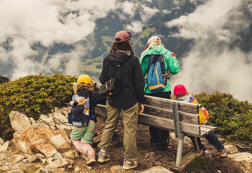 Zwei Erwachsene und zwei Kinder pausieren bei einer Bank in den Bergen und genießen die Aussicht. | © Silvretta Montafon - Roadtyping