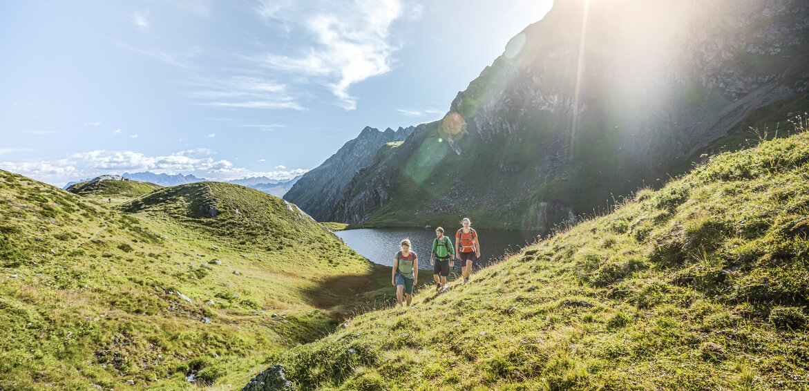 Drei Personen wandern bei Sonnenschein im Sommer in der Nähe eines Bergsees umringt von hohen Bergen. | © Silvretta Montafon - Stefan Kothner