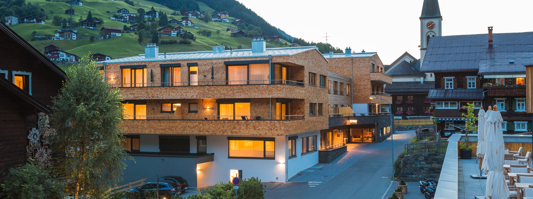 Die Chalets vom Sporthotel Silvretta Montafon und die Stoba 7 leuchten in warmen Licht mit der Kirche und den Bergen im Hintergrund in der Abenddämmerung | © Silvretta Montafon