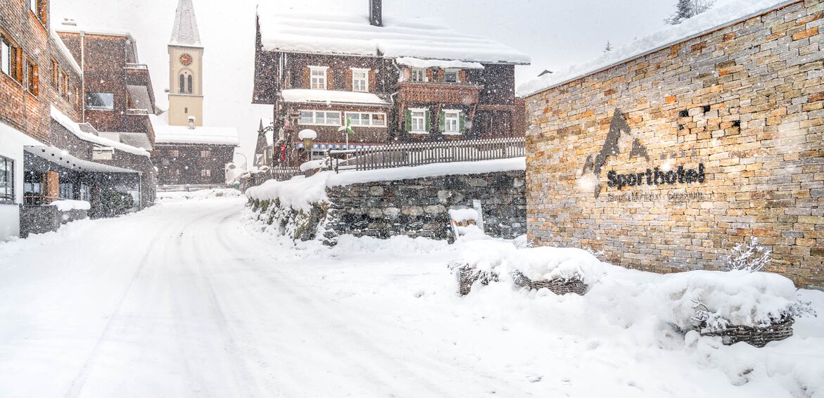 Das Sporthotel Silvretta Montafon in Gaschurn im Winter | © Silvretta Montafon - blateral Eine mit schneebedeckte Straße führt in den Dorfkern von Gaschurn mit Ausblick auf die Kirche und rechts das Sporthotel Silvretta Montafon Logo auf der Wand | © Silvretta Montafon - blateral