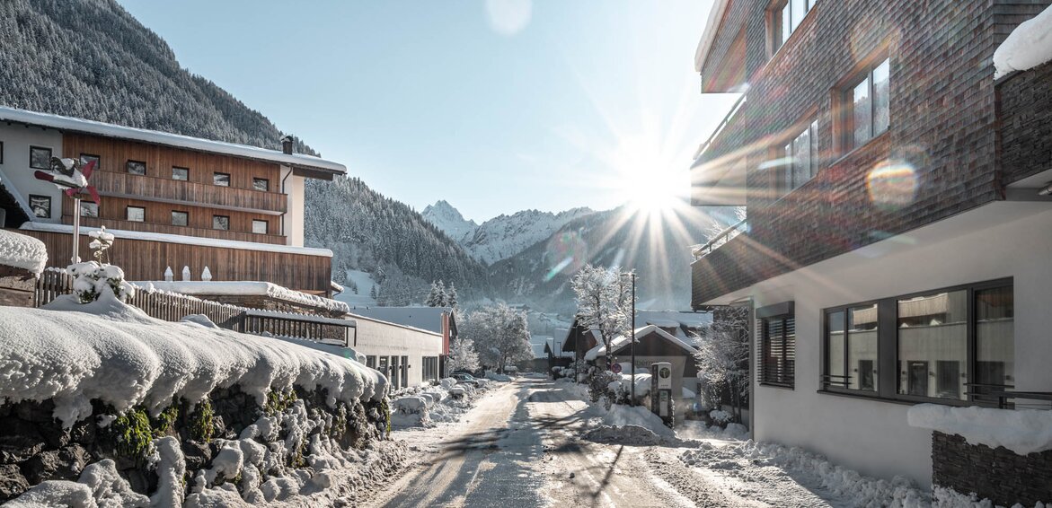 Das Sporthotel Silvretta Montafon in Gaschurn im Winter | © Silvretta Montafon - blateral Die Straße mit dem Sporthotel Silvretta Montafon links und den Chalets und der Stoba 7 rechts mit Sonne im Hintergrund und Schnee | © Silvretta Montafon - blateral