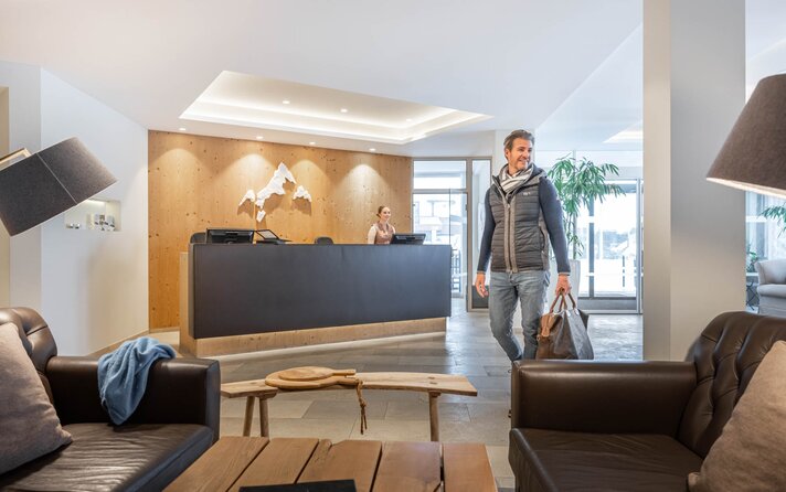 Ein Mann läuft im Eingangsbereich mit seinem Gepäck an der Rezeption vorbei in Richtung der Lounge | © Silvretta Montafon - blateral