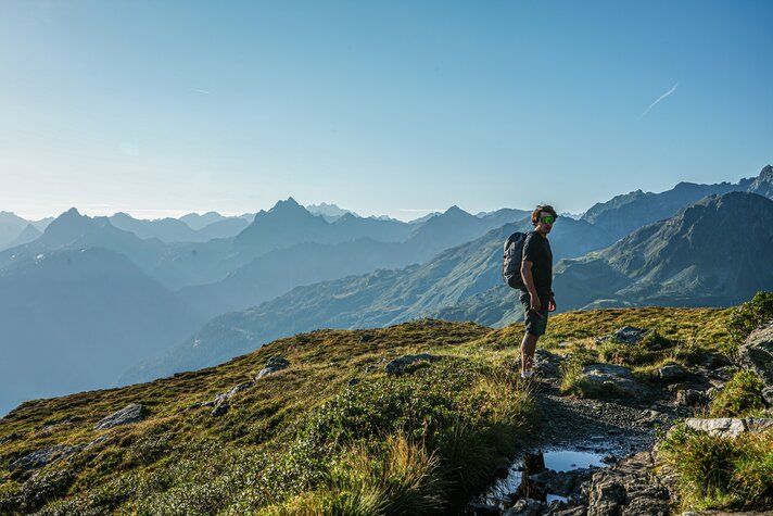 Ein Mann wandert in den Bergen im Sommer mit Aussicht auf die umliegenden Gipfel. | © Silvretta Montafon - Vanessa Strauch
