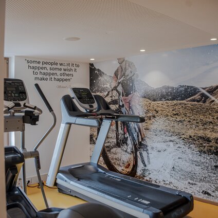 Fitnessraum mit einem Laufband im Vordergrund und einem großen Bild von einem Fahrradfahrer in den Alpen im Hintergrund | © Silvretta Montafon - Mara Baues