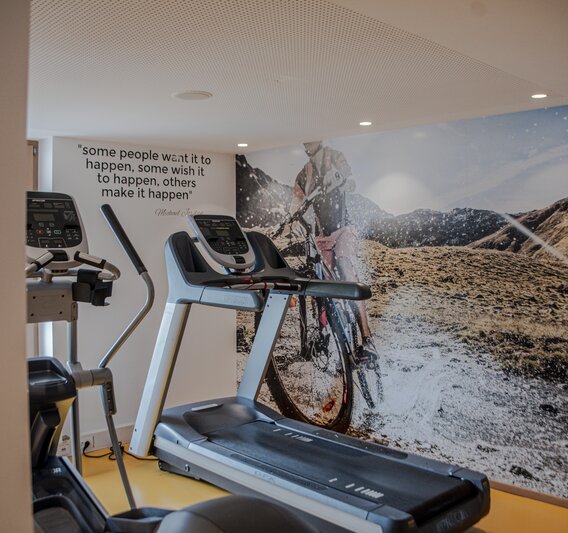 Fitnessraum mit einem Laufband im Vordergrund und einem großen Bild von einem Fahrradfahrer in den Alpen im Hintergrund | © Silvretta Montafon - Mara Baues