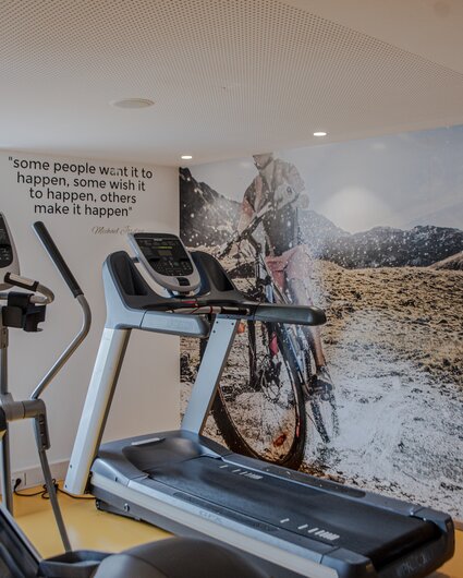 Fitnessraum mit einem Laufband im Vordergrund und einem großen Bild von einem Fahrradfahrer in den Alpen im Hintergrund | © Silvretta Montafon - Mara Baues