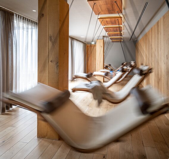 Bewegende Schwebeliegen mit einem weißen Polster in einem Raum mit Holzoptik und großer Fensterfront | © Silvretta Montafon - blateral