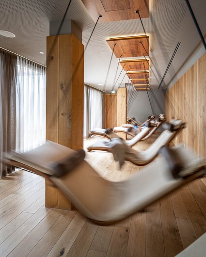 Bewegende Schwebeliegen mit einem weißen Polster in einem Raum mit Holzoptik und großer Fensterfront | © Silvretta Montafon - blateral
