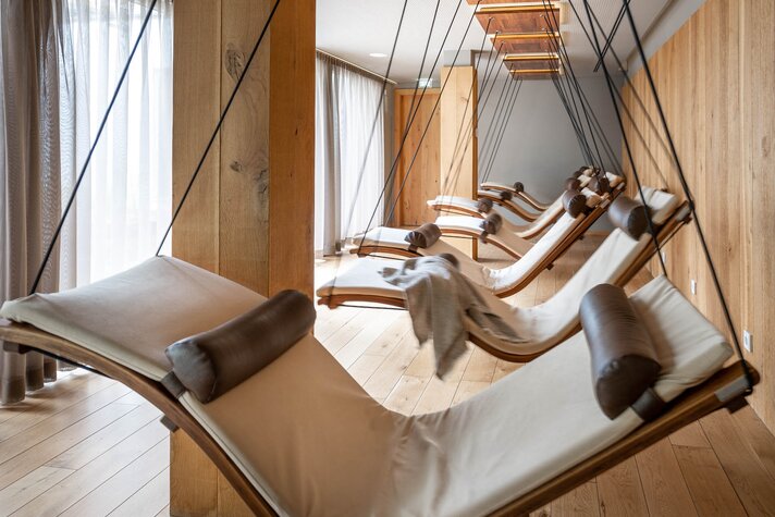 Bewegende Schwebeliegen mit weißer Polsterung in einem Raum mit Holzoptik und einer Fensterfront | © Silvretta Montafon - blateral