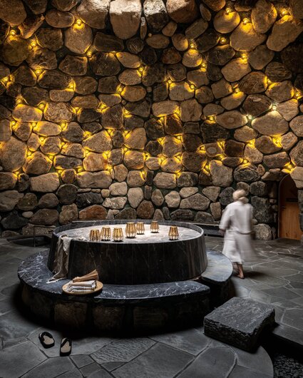 Eine Frau läuft durch das Tepidarium, welches in goldenes Licht getaucht ist mit einem Loch in der Decke.  | © Silvretta Montafon - blateral