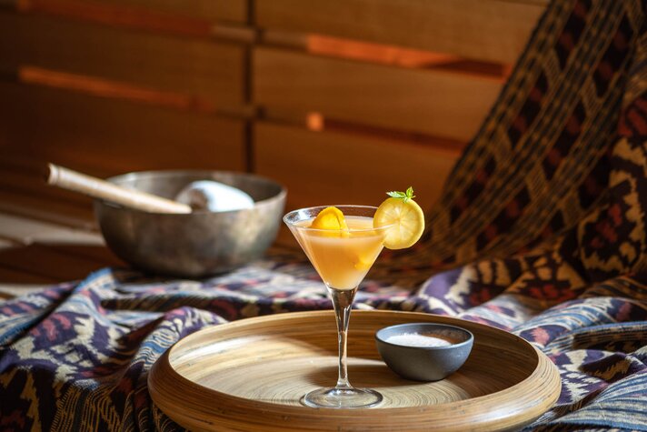 Gold Spa im Sporthotel Silvretta Montafon | © Silvretta Montafon - blateral Ein gelber Cocktail mit Zitronenscheibe auf einem Holztablett mit einem Schälchen Salz und einer Klangschale  | © Silvretta Montafon - blateral