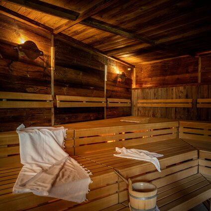 Eine finnische Sauna mit Holzwänden und Holzbänken, auf welchem ein weißes Handtuch liegt und ein Wasserkübel aus Holz steht | © Silvretta Montafon - blateral