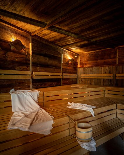 Eine finnische Sauna mit Holzwänden und Holzbänken, auf welchem ein weißes Handtuch liegt und ein Wasserkübel aus Holz steht | © Silvretta Montafon - blateral