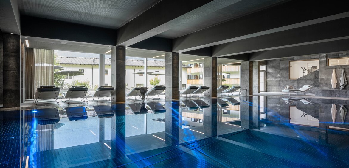 Der blaue Kristallpool im Inneren vom Sporthotel Silvretta Montafon mit Liegen und einer großen Fensterfront mit Blick ins Grüne | © Silvretta Montafon - blateral