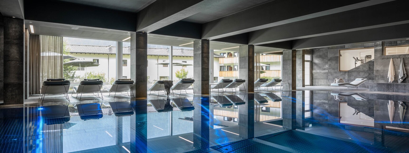 Kristallpool im Sporthotel Silvretta Montafon | © Silvretta Montafon - blateral Der blaue Kristallpool im Inneren vom Sporthotel Silvretta Montafon mit Liegen und einer großen Fensterfront mit Blick ins Grüne | © Silvretta Montafon - blateral