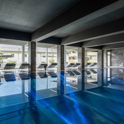 Der blaue Kristallpool im Inneren vom Sporthotel Silvretta Montafon mit Liegen und einer großen Fensterfront mit Blick ins Grüne | © Silvretta Montafon - blateral