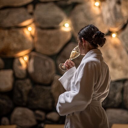 Eine Frau in einem Bademantel trinkt einen Prossecco im Tepidarium mit goldenem Licht in den Steinwänden | © Silvretta Montafon - Mara Baues