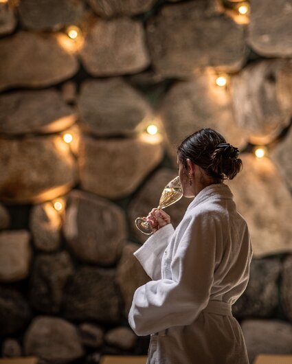 Eine Frau in einem Bademantel trinkt einen Prossecco im Tepidarium mit goldenem Licht in den Steinwänden | © Silvretta Montafon - Mara Baues
