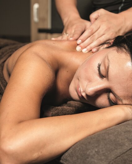 Eine dunkelhaarige Frau liegt auf einer Massageliege und bekommt eine Rückenmassage | © Silvretta Montafon
