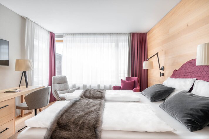 Doppelzimmer im Sporthotel Silvretta Montafon | © Silvretta Montafon - blateral Ein gemütliches Doppelbett mit weinroter Polsterung und weißem Bettbezug | © Silvretta Montafon - blateral