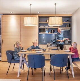 Drei Kinder sitzen gemeinsam am Tisch währenddessen der Vater am kochen ist | © Silvretta Montafon - blateral