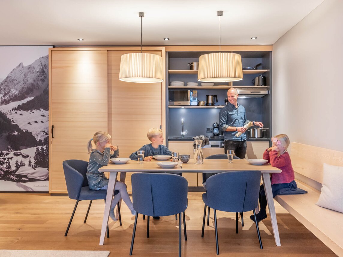 Drei Kinder sitzen gemeinsam am Tisch währenddessen der Vater am kochen ist | © Silvretta Montafon - blateral