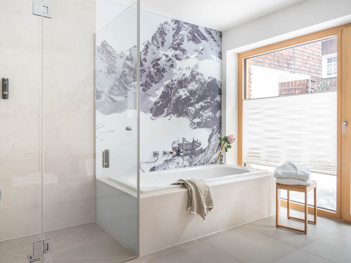 Eine geräumige ebenerdige Dusche, eine Badewanne mit einem Skibild im Hintergrund einer großen Glasfront | © Silvretta Montafon - blateral