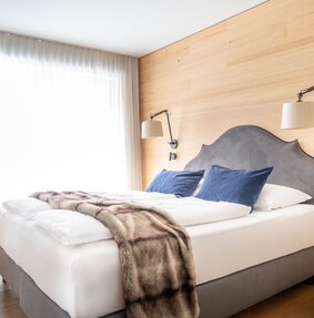 Ein großes Bett mit grauer Polsterung, einer weißen Decke und einem blauen Vorhang im Hintergrund | © Silvretta Montafon - blateral